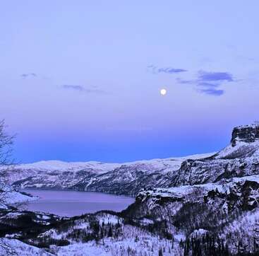 Uma paisagem serena de inverno apresenta montanhas e árvores cobertas de neve, um rio ou lago tranquilo e um céu encantador, com a lua cheia iluminando a cena de forma linda.