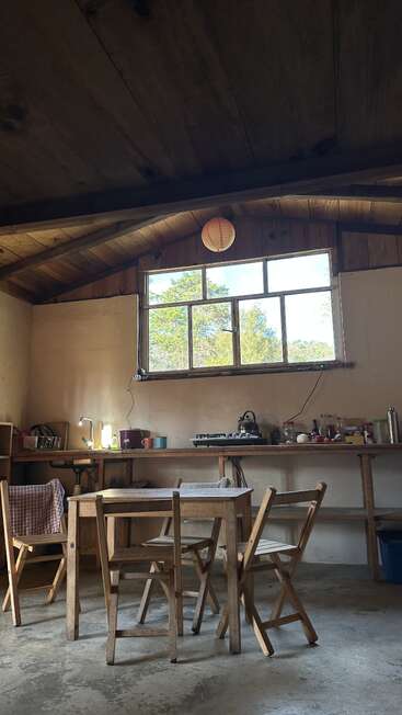 Une cuisine rustique avec des meubles en bois, une grande fenêtre, un aménagement simple et beaucoup de lumière naturelle. On y trouve quatre chaises, une table et quelques ustensiles de cuisine sur le plan de travail.