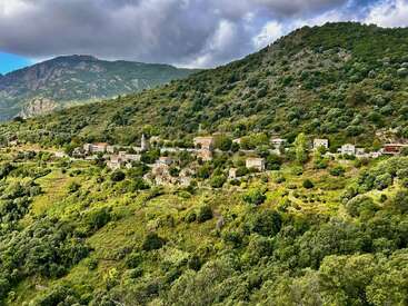 This image shows a small village nestled among green hills and dense forest, with mountains in the background under dramatic, cloudy skies, conveying peaceful seclusion.