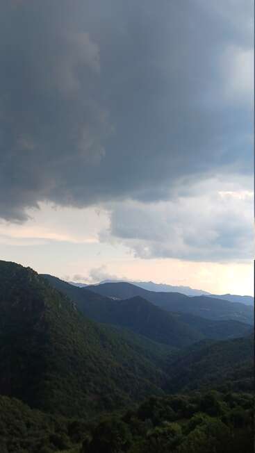 Dark, heavy clouds loom over lush, green mountains. The landscape features deep valleys, distant peaks, and a soft haze, creating a dramatic, moody, and peaceful atmosphere.