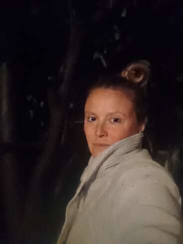A woman stands outside at night, wearing a light-colored jacket with a high collar. Her hair is up in a bun. Background is dark and shadowy.