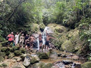 Eine Gruppe von Menschen und ein Hund posieren fröhlich vor einem malerischen Wasserfall, umgeben von üppig grünem Wald und moosbewachsenen Felsen, und genießen gemeinsam die Natur.
