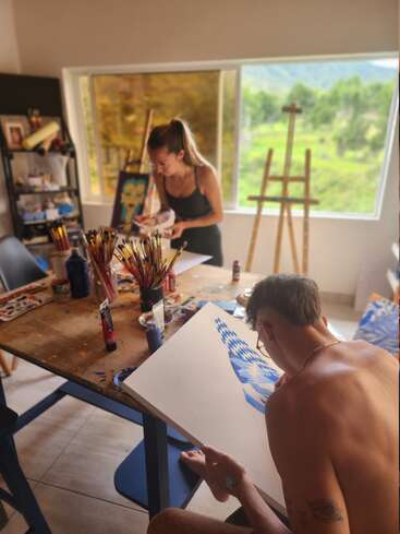 Zwei Menschen malen in einem hellen, mit Kunst gefüllten Atelier mit großen Fenstern und Blick ins Grüne. Malutensilien, Leinwände und Pinsel bedecken den Holztisch und schaffen eine kreative Atmosphäre.
