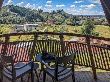 Ein gemütlicher Balkon mit zwei Stühlen und einem Tisch bietet einen Blick auf eine malerische Landschaft mit Hügeln, Grünflächen, verstreuten Häusern und einem blauen Himmel mit flauschigen Wolken.