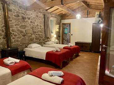 This cozy rustic bedroom features three single beds with red blankets, stone walls, wooden ceiling beams, a wood stove, wardrobe, and soft, warm lighting.
