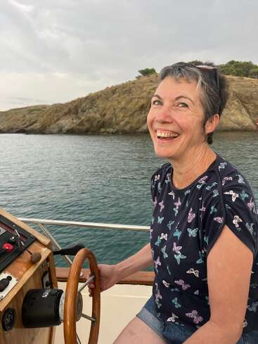 Eine fröhliche Frau mit kurzen Haaren und einem Hemd mit Schmetterlingsmuster steuert ein Boot. Sie lächelt breit, umgeben von ruhigem Wasser und felsigen Ufern.