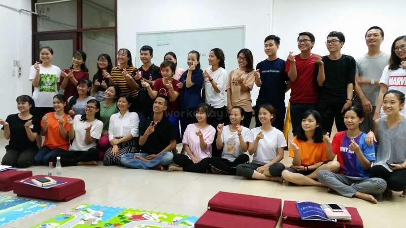 The image shows a group of young people posing for a photo, sitting on the floor with their hands in a gesture, in a room with white walls and a map on the floor.