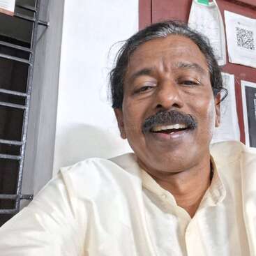 Un hombre sonriente, con bigote y vestido con una camisa de color claro, se hace un selfi en el interior de una habitación. Detrás de él hay papeles, un calendario y una reja metálica en la ventana.