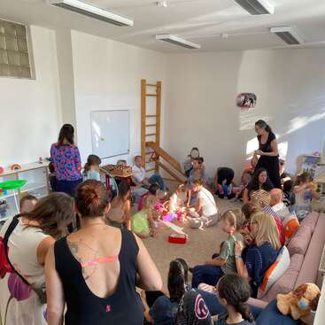 Uma sala bem iluminada, repleta de crianças e adultos envolvidos em várias atividades, brincando, conversando e sentados juntos em sofás e no piso acarpetado. Atmosfera animada.
