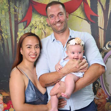 A smiling family poses together. The woman is in a blue dress, the man holds a happy baby with a floral headband. Colorful, festive background.