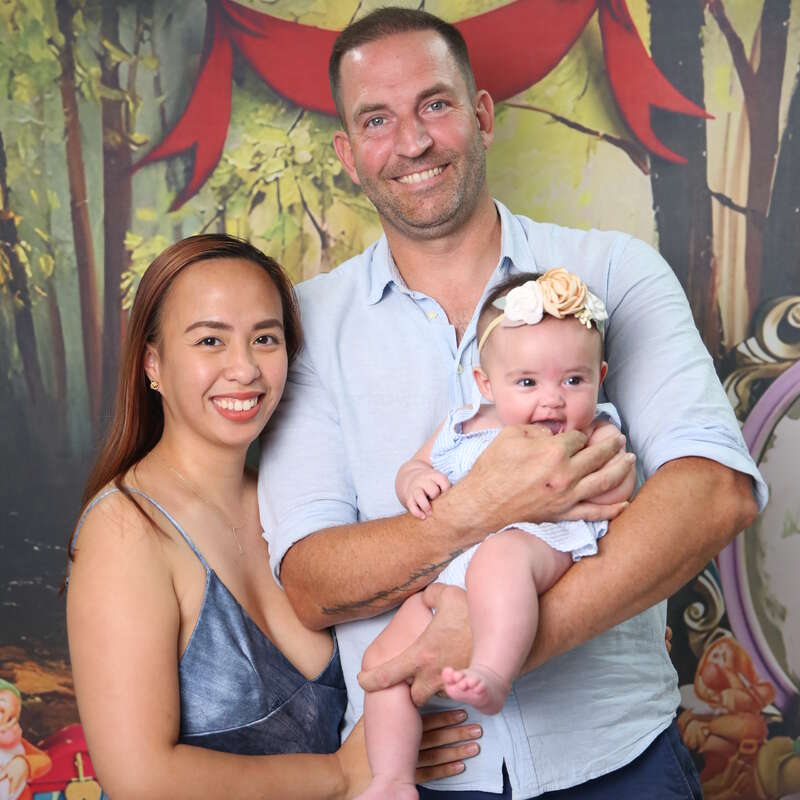 Uma família sorridente posa junta. A mulher está com um vestido azul e o homem segura um bebê feliz com uma faixa floral na cabeça. Fundo colorido e festivo.