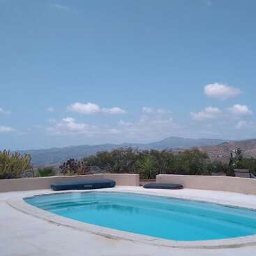 Ein ruhiger Swimmingpool mit klarem, blauem Wasser, umgeben von Wüstenvegetation und einer niedrigen Mauer, mit Blick auf die fernen Berge unter einem hellen, teilweise bewölkten Himmel. Friedliche Atmosphäre.