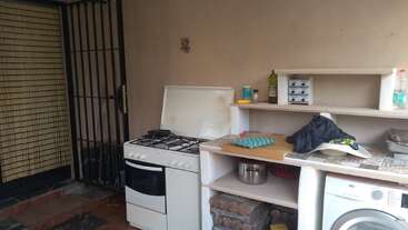 Dieses Bild zeigt eine kleine Küchenecke mit einem Gasherd, offenen Regalen mit Töpfen, Eiern und Küchenutensilien sowie einer Waschmaschine und einigen verstreuten Utensilien.