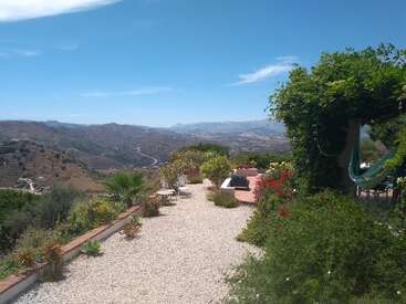 Eine wunderschöne Gartenterrasse mit Blick auf sanfte Hügel und Berge unter einem blauen Himmel. Hängematte, Pflanzen, Blumen und Gartenmöbel laden zum Entspannen in diesem malerischen Paradies ein.