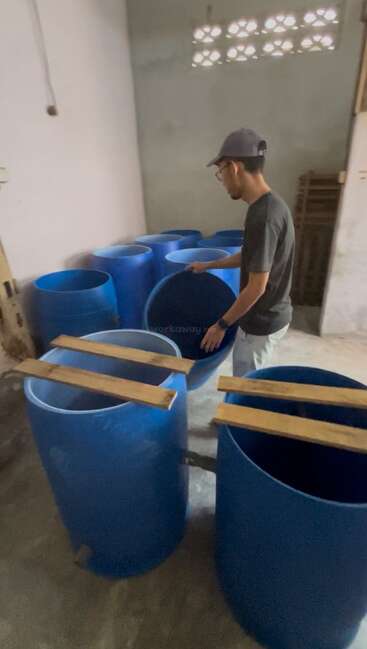 Una persona con gorra y gafas está de pie en una habitación llena de grandes barriles de plástico azul y tablones de madera, posiblemente preparando artículos para almacenaje o construcción.