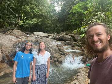 Tres amigos sonríen frente a un arroyo rocoso y una pequeña cascada rodeados de un frondoso bosque verde, disfrutando de un día al aire libre y del refrescante paisaje natural.