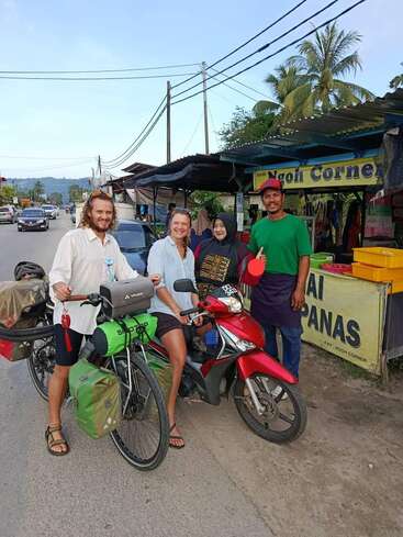 Cuatro personas sonrientes posan delante de un puesto de comida al borde de la carretera, con dos bicicletas cargadas de material de viaje. Al fondo, palmeras y tiendas bajo un cielo azul.