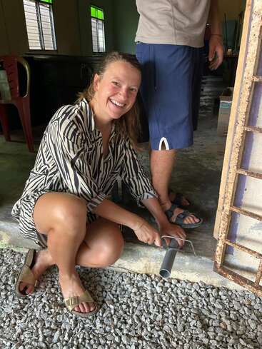 Una mujer sonriente con camisa de rayas se agacha en el suelo, utilizando una sierra para cortar una tubería de metal junto a un hombre que está de pie cerca, en el interior.