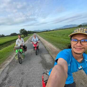 Tres amigos disfrutan de una aventura al aire libre en una carretera rural: uno en bicicleta, otro en scooter y otro tomándose un selfie mientras conducen.