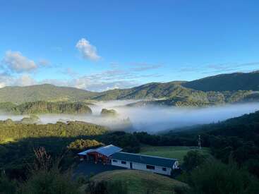 Uma casa fica tranquila em um vale verdejante, cercada por colinas e árvores enevoadas sob um céu azul claro com nuvens dispersas. Uma manhã serena.