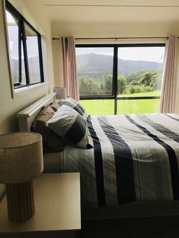 Esse quarto aconchegante tem uma colcha listrada, mesa de cabeceira com abajur, janelas grandes e cortinas cor-de-rosa, com vista para colinas verdejantes e uma bela vista panorâmica.