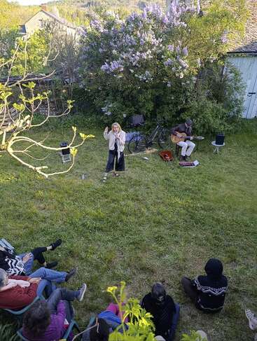 In einem üppigen Garten findet ein kleines Open-Air-Konzert statt. Eine Frau singt, begleitet von einem Gitarristen, während sich das Publikum entspannt auf dem Rasen sitzt.