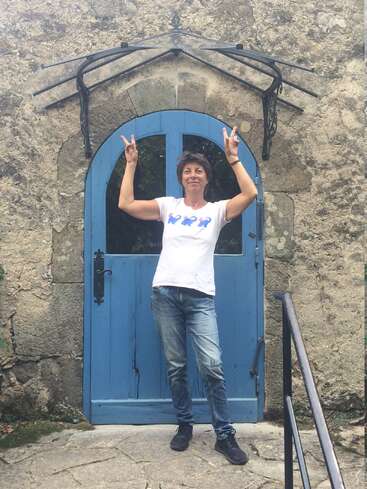 Eine Frau steht vor einem rustikalen Steinhaus mit einer blauen Bogentür, macht mit beiden Händen Peace-Zeichen, lächelt selbstbewusst und trägt Jeans und ein weißes T-Shirt.