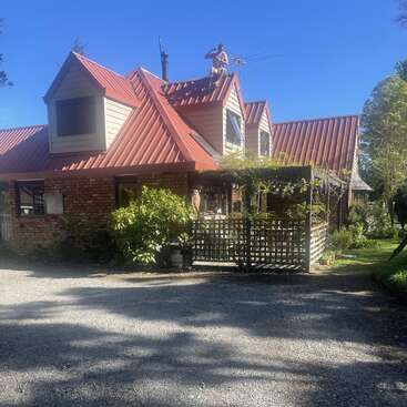 A brick house with a red metal roof and dormer windows. A person is fixing or inspecting an antenna on the roof under a clear blue sky.