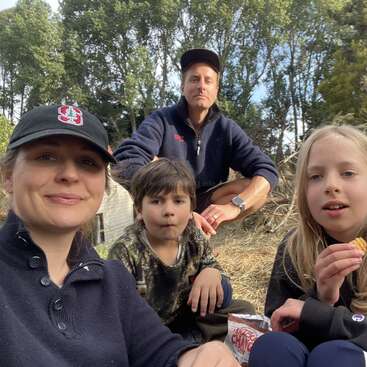 Una familia de cuatro miembros está sentada al aire libre, disfrutando de unos aperitivos y de la naturaleza. Parecen relajados y felices, rodeados de árboles y vegetación, compartiendo un momento de tranquilidad juntos.