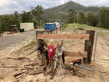 The image depicts two young girls posing with two dogs in front of a wooden fence, set against a backdrop of rolling hills and trees.