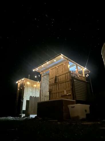A rustic wooden structure with corrugated metal roof is illuminated by string lights at night, under a starry sky. A white storage box sits outside.