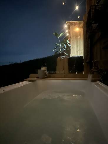 A cozy outdoor bathtub filled with water, surrounded by warm string lights, a potted plant, and wooden walls, creating a relaxing and tranquil nighttime atmosphere.