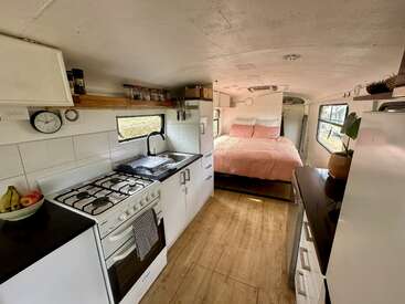 This image shows a cozy, converted bus interior featuring a compact kitchen with a stove and sink, stylish white cabinets, wood flooring, and a inviting bed.