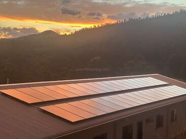 Solar panels are installed on a rooftop, reflecting the warm colors of a beautiful sunset sky. Forested hills create a scenic, tranquil background.