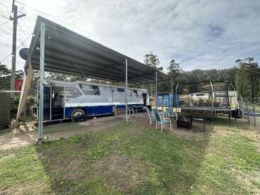 A converted bus sits under a metal carport beside patio chairs and a trampoline, surrounded by trees, an open yard, and a partly cloudy sky.