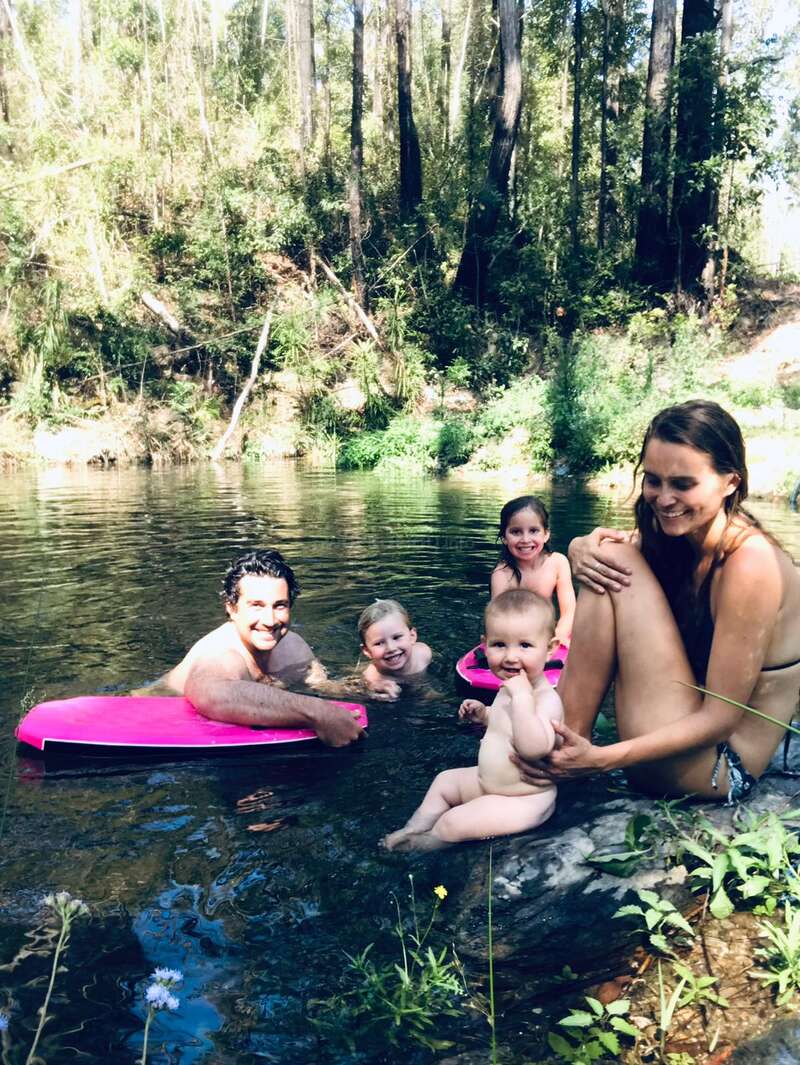 The image depicts a family of five enjoying a day at the river, with two adults and three children, all smiling and posing for the camera in the water.