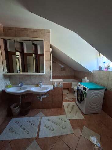 Esse banheiro tem teto inclinado, azulejos marrons, pias duplas, um espelho grande, máquina de lavar roupa, banheira de canto, vaso sanitário e vários tapetes brancos com desenhos de peixes.