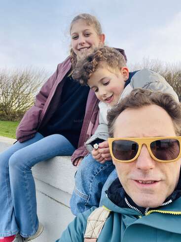 Un hombre con gafas de sol se hace una alegre selfie al aire libre junto a dos niños sonrientes sentados a su lado sobre un muro blanco, disfrutando de un día soleado y tranquilo.