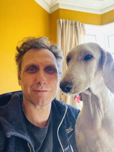 Un hombre sonríe a la cámara mientras está sentado junto a un perro blanco en una habitación muy iluminada, con paredes amarillas y cortinas de color crema junto a la ventana.
