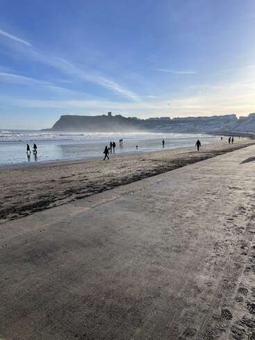 Una preciosa escena de playa con gente paseando por la orilla bajo la luz del sol, acantilados en la lejanía, olas suaves y un cielo azul brillante en lo alto. Ambiente tranquilo.