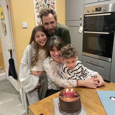 Una familia se reúne en una acogedora cocina, sonriendo alrededor de una tarta de chocolate con velas encendidas que forman el «43», celebrando juntos un alegre cumpleaños lleno de amor y felicidad.
