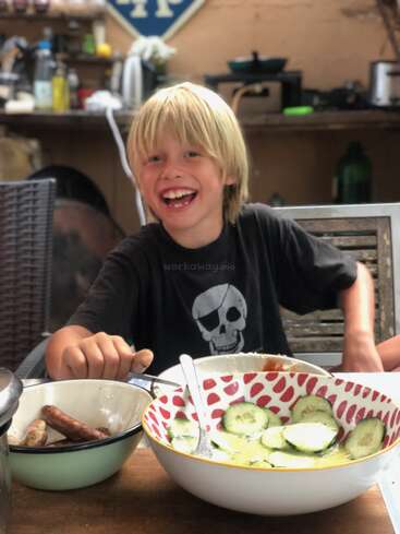 Un chico rubio y sonriente se sienta a una mesa con cuencos de comida, que incluyen salchichas y ensalada de pepino, disfrutando de una comida en un ambiente acogedor, parecido a una cocina.