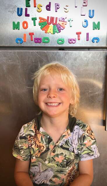 Un niño rubio sonriente está de pie delante de una nevera metálica decorada con letras magnéticas de colores. El niño lleva una camiseta con motivos de tigres y follaje.