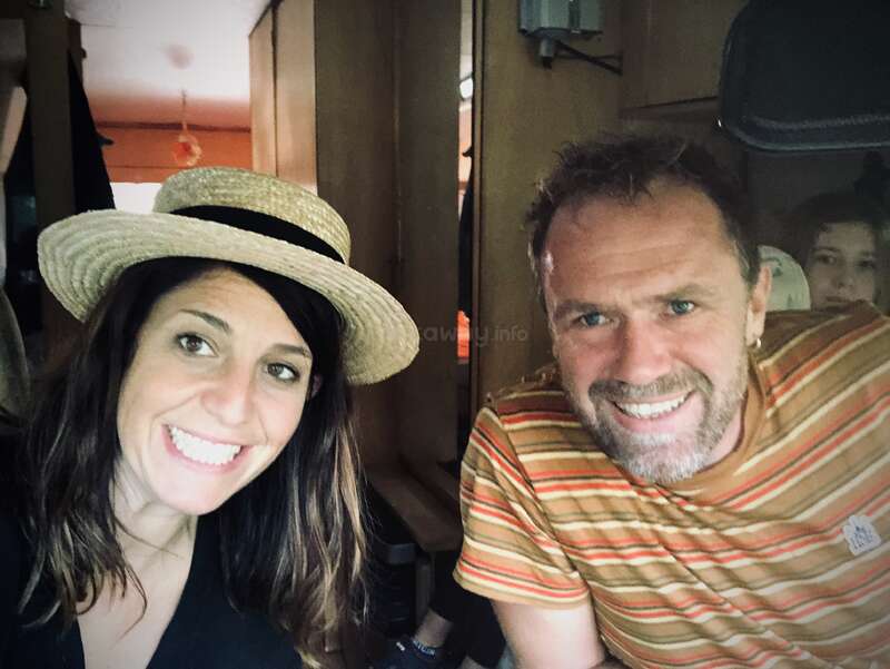 La imagen muestra un selfie de una mujer y un hombre sonriendo a la cámara, con la mujer con sombrero de paja y el hombre con camisa de rayas.