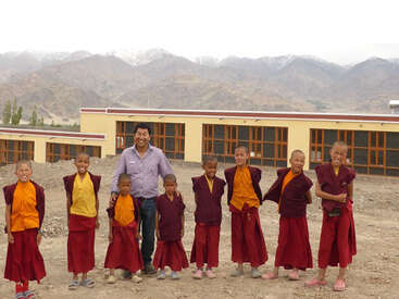 The image depicts a group of young Buddhist monks standing in front of a building, with a man in a light-colored shirt and dark pants posing with them.