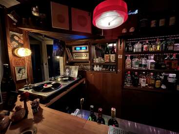 The image depicts a dimly lit bar with a turntable, bottles of alcohol, and a shelf of liquor, creating a cozy and intimate atmosphere.