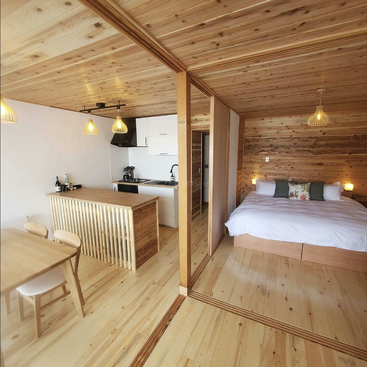 This cozy studio apartment features wood-paneled ceilings and floors, a compact kitchen, dining area, and a comfortable double bed, all separated by a sliding glass partition.
