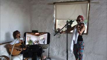 Dois músicos se apresentam em uma sala aconchegante e rústica. Um toca violão sentado, o outro toca violino em pé. Um smartphone grava a sessão perto de uma janela iluminada.