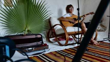 Uma pessoa senta-se em uma cadeira de vime tocando violão, cercada por equipamentos musicais. Folhas de palmeira, um estojo de violão e um tapete listrado completam a cena aconchegante.
