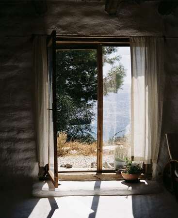 A luz do sol entra por uma porta aberta com cortinas transparentes, revelando uma vista serena de árvores, água azul e céu do lado de fora de um cômodo rústico e tranquilo.